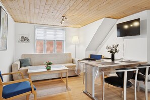 Una sala moderna con techo de madera, un sofá, una mesa de centro, una mesa de comedor con sillas, un televisor montado en la pared y una ventana con persianas.