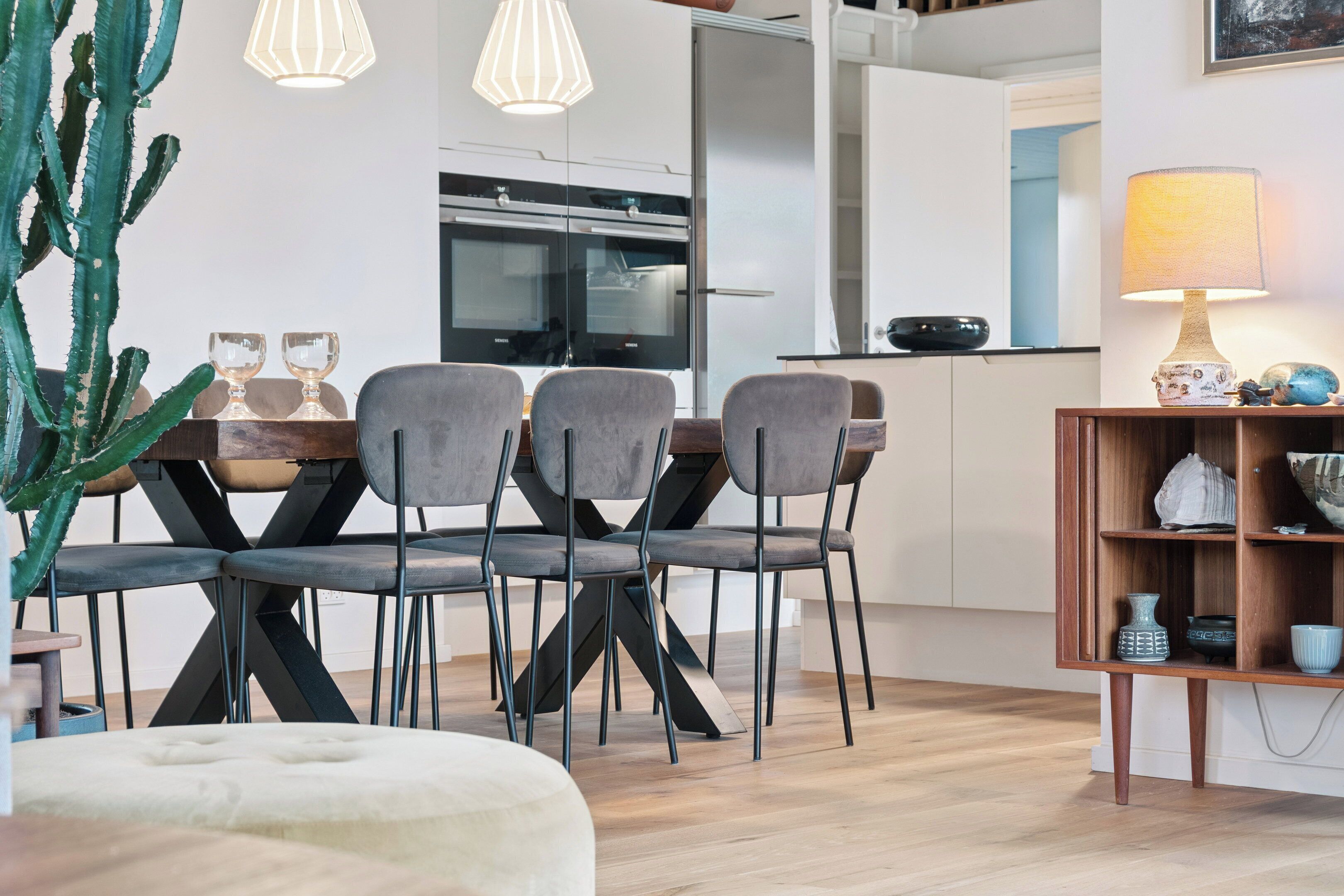 A modern kitchen with a dining area, featuring a cactus plant, dining chairs, a table, pendant lights, and a lamp.