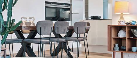 A modern kitchen with a dining area, featuring a cactus plant, dining chairs, a table, pendant lights, and a lamp.