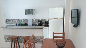 Eine kleine Küche mit Mikrowelle, Kaffeemaschine und Kühlschrank. Zwei hölzerne Hocker und ein Tisch mit einer einzelnen Schüssel sind vorhanden.