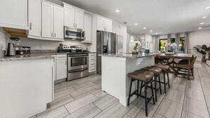 A modern kitchen with white cabinets, stainless steel appliances, a central island with stools, and a dining area with a table and chairs. - 8 Br Magical Themed Villa Close To Disney (Kissimmee)