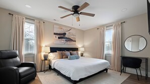 A modern bedroom with a large bed, a ceiling fan, a black leather chair, a desk, and a round mirror.