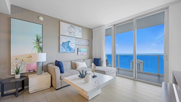 A modern living room with a sofa, coffee table, and artwork. A large window offers a view of the ocean. - Luxury 32nd Floor Condo | Full Beach Club Access (Hollywood)