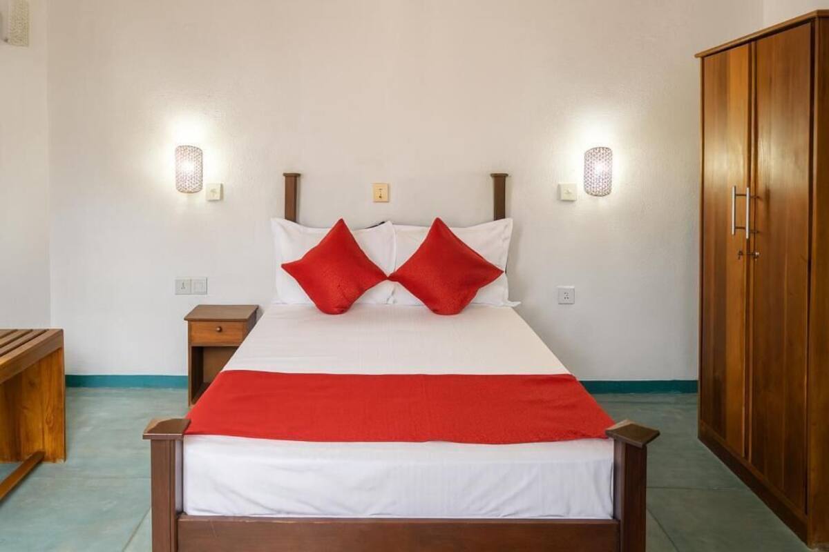 Un letto rifatto con una coperta rossa e bianca, due cuscini rossi e una testiera in legno. Si notano due comodini, uno con una lampada, e un'armadio in legno sulla destra.