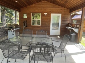 A covered outdoor area with a metal table and chairs, a grill, and a wooden cabin structure.
