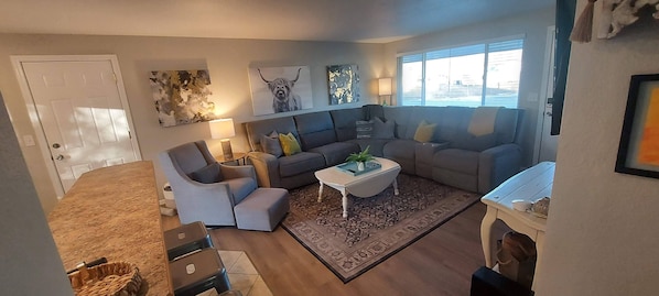 A living room with a sofa, armchairs, a coffee table, and a rug. There is a door, a lamp, and artwork on the walls.