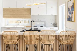 Une cuisine moderne avec des armoires blanches, une cuisinière encastrée et une fenêtre au-dessus du comptoir. On y trouve quatre chaises en osier près du comptoir.