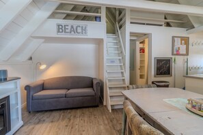 Une pièce chaleureuse avec un canapé à carreaux gris, une table à manger en bois, un escalier portant l’inscription « PLAGE » et une petite cheminée.