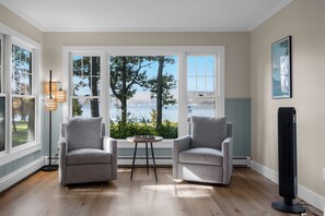 Une pièce avec deux fauteuils gris, une petite table et un ventilateur sur pied. De grandes fenêtres offrent une vue sur des arbres et une étendue d’eau.