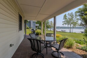 A covered patio with a glass table and chairs, overlooking a lake with a grassy area and trees. - Lovely Views of Lake Leelanau at Bluebird Landing (Cedar)