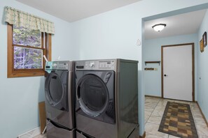 A laundry room with two front-loading washing machines, a window with a view of trees, and a door leading to another room. - Ideal Suttons Bay Retreat at Broadway Base Camp (Suttons Bay)
