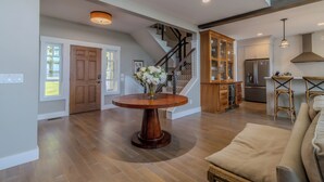 A spacious living area with a wooden floor, a round wooden coffee table, a beige sofa, a kitchen with a refrigerator, and a staircase with wooden railings. - Dreamy Northern Michigan Escape, Chateau Sunrise (Suttons Bay)
