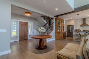 A spacious living area with a wooden floor, a round wooden coffee table, a beige sofa, a kitchen with a refrigerator, and a staircase with wooden railings. - Dreamy Northern Michigan Escape, Chateau Sunrise (Suttons Bay)