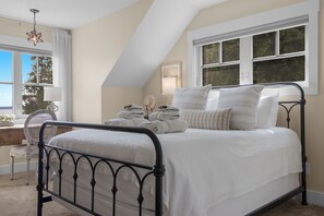 Un dormitorio con cama, almohadas, mesita de noche, lámpara y ventana con vistas a árboles.