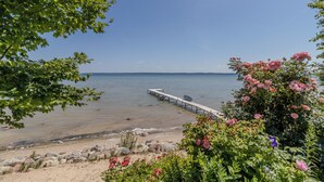 A beach with a pier extending into calm waters, surrounded by lush greenery and flowering plants.