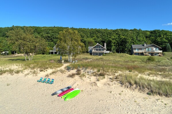 Ein Strandgrundstück mit einem großen Haus, einer mit Liegestühlen ausgestatteten Strandpromenade und einer bewaldeten Hügelkette im Hintergrund.