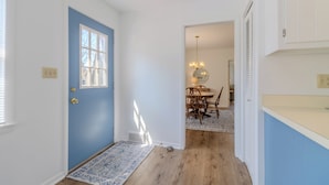 Um corredor com uma porta azul, piso de madeira, uma sala de jantar com lustre e uma cozinha com armários brancos.