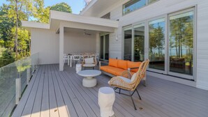 A modern outdoor deck with a glass balustrade, white furniture, and a view of the surrounding trees.