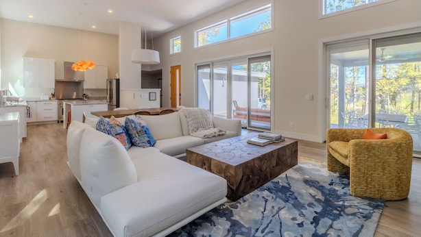 A spacious living room with a white sofa, a wooden coffee table, and a yellow armchair. There is a kitchen in the background with white cabinets and a hanging light fixture.
