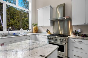 A modern kitchen with a large marble countertop, stainless steel appliances, and a view of trees through the window. - Stargazer – Modern Luxury Meets Up North Charm (Northport)