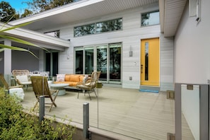 A modern outdoor patio with a wooden deck, a yellow door, and a seating area with wicker chairs and a sofa. - Stargazer – Modern Luxury Meets Up North Charm (Northport)