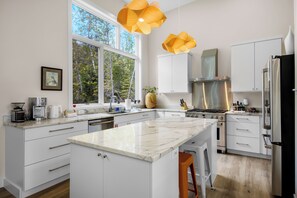 Una cocina moderna con una isla grande, muebles blancos y vistas a árboles por la ventana.