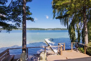 A wooden deck with a bench and chairs overlooking a calm lake with a pier extending into the water.
