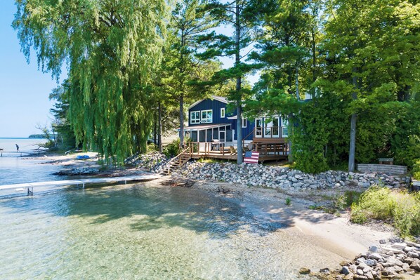 A lakeside cabin with a deck, surrounded by trees and a rocky shoreline.