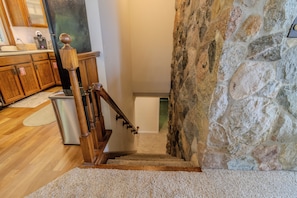 A staircase with wooden railings leading to a lower level, a stone wall on the right, and a kitchen area with wooden cabinets and a stainless steel appliance.
