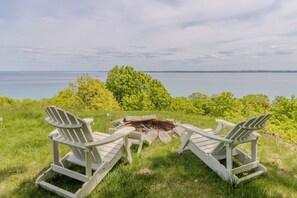 Two Adirondack chairs facing a fire pit with a view of a lake and trees in the background.