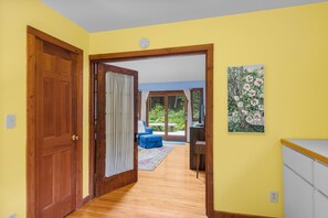 Un pasillo con puertas de madera, un cuadro de flores y vista a una habitación con pared azul y una ventana.