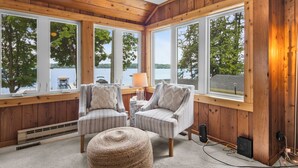 A room with wooden paneled walls, large windows, two upholstered chairs, a small round ottoman, and a lamp on a side table. - Cozy Cottage on Long Lake, Your Perfect TC Getaway (Traverse City)
