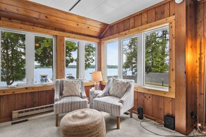 Ein Zimmer mit Holzvertäfelung, großen Fenstern, zwei Polsterstühlen, einem kleinen runden Hocker und einer Lampe auf einem Beistelltisch.