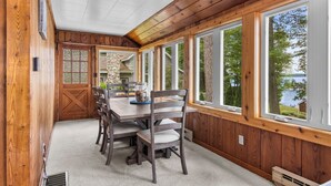 A wooden-paneled room with a dining table, chairs, and large windows overlooking a lake. - Cozy Cottage on Long Lake, Your Perfect TC Getaway (Traverse City)