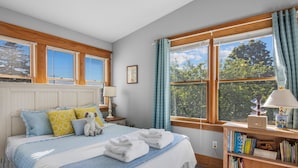 Un dormitorio con cama, almohadas, una mesita de noche con lámpara, una estantería y una ventana con cortinas.