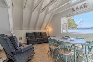 Una habitación acogedora con sofá, sillón y mesa de comedor con sillas. Se ve una ventana con vistas a un lago.