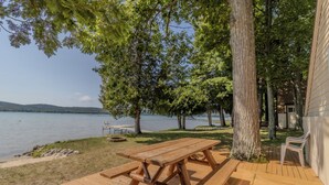 Une terrasse en bois avec des tables de pique-nique et des bancs, donnant sur un lac bordé d’arbres, avec un bâtiment visible au loin.
