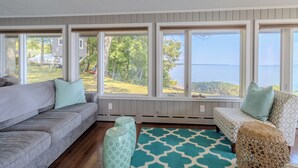A room with a gray sofa, a patterned ottoman, and a view of a lake through large windows. - Panoramic Point - dazzling views and private beach (Traverse City)