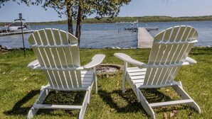 Twee witte Adirondack stoelen gericht op een waterlichaam met een steiger en een boom op de achtergrond.