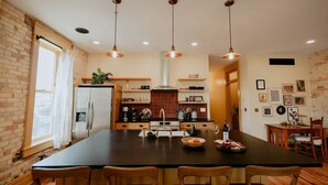 A modern kitchen with a large island, pendant lights, and a brick wall.