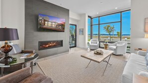 A modern living room with a fireplace, a flat-screen TV, and a large window offering a view of the outdoors.