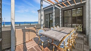 A covered outdoor dining area with a wooden table and wicker chairs, overlooking a beach.