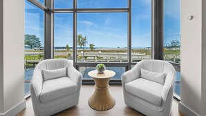 A room with a large window, two white armchairs, a small round table with a plant, and a view of a body of water and distant landscape.