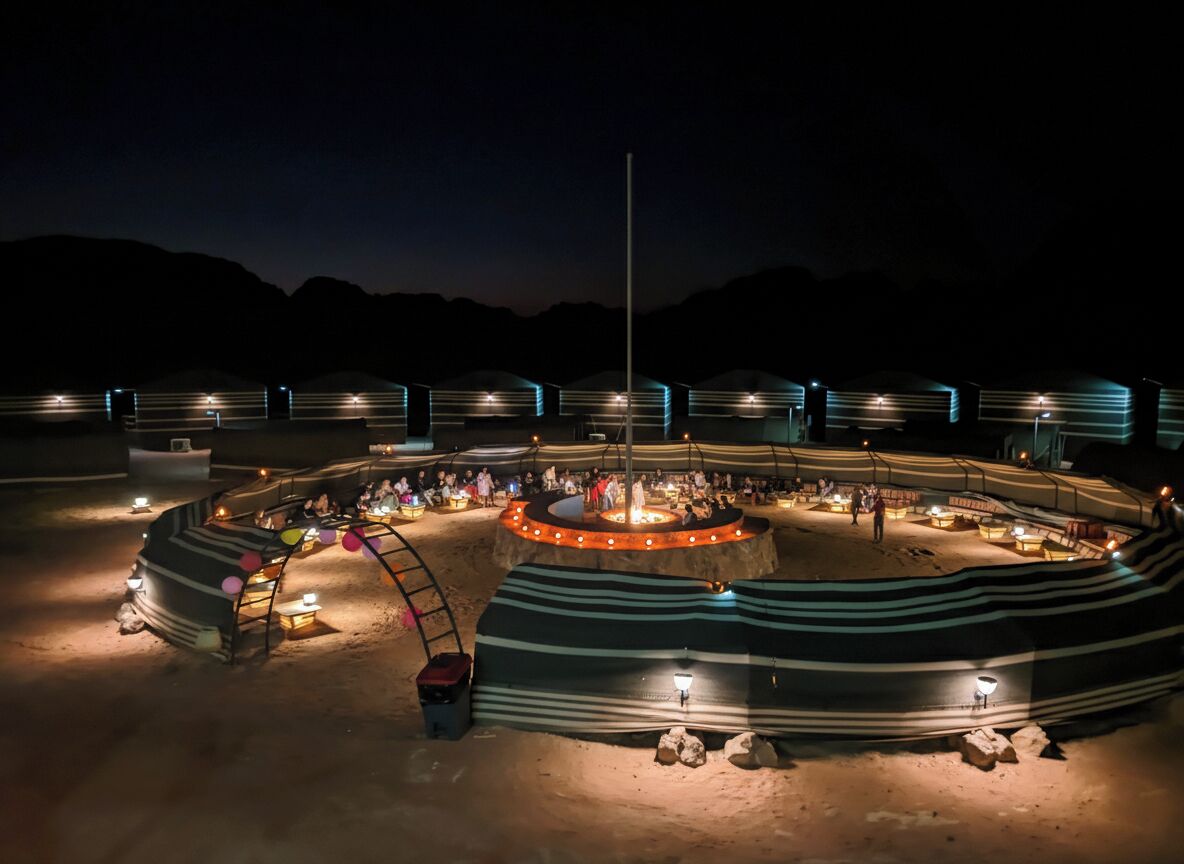 A circular outdoor structure with multiple tents and a central fire pit, surrounded by a sandy area with rocks and a flagpole.