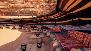 An outdoor seating area with stone benches and tables, covered by striped fabric canopies, set against a backdrop of rock formations. - Desert Sand Camp (Wadi Rum)