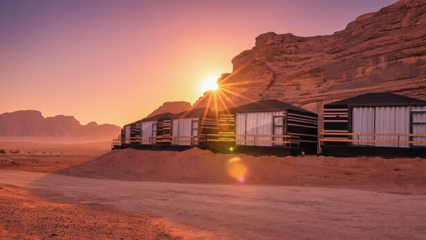 A desert landscape with a row of caravan-style tents with a dirt road in the foreground and a rocky cliff in the background. - Desert Sand Camp (Wadi Rum)