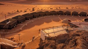 A desert landscape with tents arranged in a semi-circle, a fenced area, and a view of distant mountains at sunset. - Desert Sand Camp (Wadi Rum)