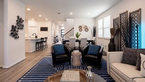 Een moderne woonkamer met een bank, twee fauteuils, een salontafel en een vloerkleed. Achterin is een keuken met een eettafel en stoelen.