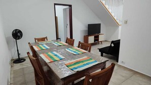 A dining area with a long table, chairs, a TV, and a fan. - Brisa e Mar Townhouse (Caraguatatuba)