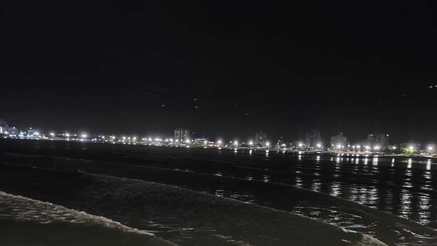 A nighttime beach scene with waves crashing onto rocks and a city skyline in the background. - Brisa e Mar Townhouse (Caraguatatuba)